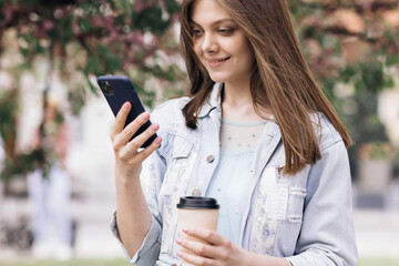 Attractive young woman with smartphone and cup of coffee in hands on background of sakura tree in city park . Pretty summer woman in white dress looking at her mobile phone and smile. Stylish Outfit © uflypro