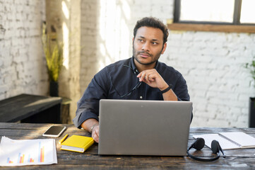 Smart and clever multiracial male entrepreneur thinks about way to develop his startup, thoughtful intelligent hindu man in smart casual wear using laptop in modern loft office space