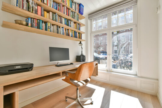 Modern interior of light study room with wooden wall mounted table and computer under shelves with books