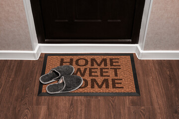 Doormat with text Home sweet home before the dark door in the hall. Home slippers on the rug. Top view of mat on wooden floor.
