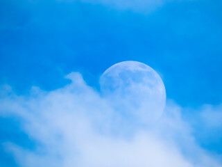 An incomplete moon in the afternoon against a blue sky partially hidden by a white cloud