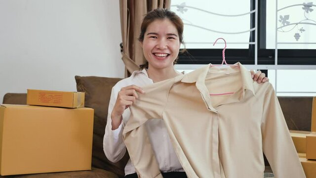 Happy Asian young woman is online small business owner sitting on couch and live streaming to online for selling clothes by smartphone at home office. Entrepreneur startup ecommerce marketing concept.