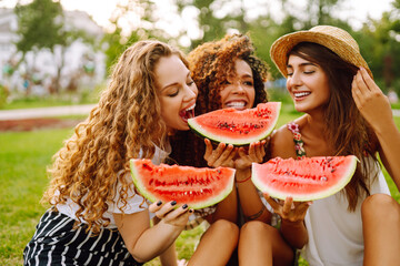 Cheerful happy friends camping on the grass, eating watermelon, laughing. Three young woman relaxing and enjoying holidays together. People, lifestyle, travel, nature and vacations concept.