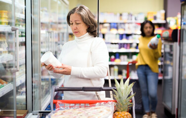 Portrait of interested elderly woman shopping in supermarket, choosing fresh dairy products on...