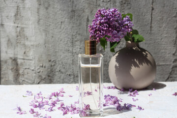  perfume bottle standing on a neutral background and surrounded by lilac flower petals. Behind in vase u see fresh lilac flower. Idea for floral scented perfume advertisement and mock up