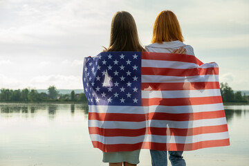 Two young friends women with USA national flag on their shoulders hugging together outdoors on lake...