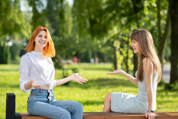 Two young women friends sitting on a bench in summer park and talking having an argument.