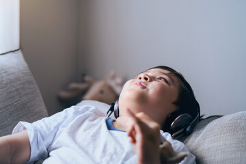 Close up face of happy kid listens to music on headphones, Candid shot cute boy looking up with smiling face, Child lying on sofa wearing earphones listening relaxing music. Positive children concept