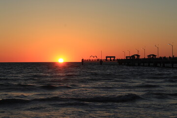 Naklejka na ściany i meble Fishing pier at the beach with the sun on the horizon at sunset