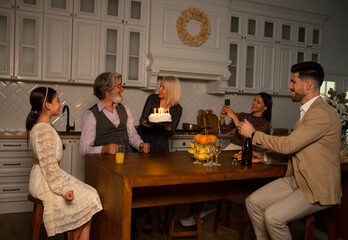 Making wish. Big happy multigenerational family celebrating birthday of grandfather sitting at table in kitchen at home in evening. Grandmother bringing cake with candles while smiling at her husband