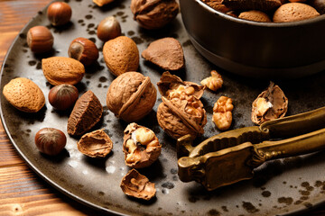 Almonds, walnuts and hazelnuts are sitting in a brown bowl and a nutcracker on a dark brown spotted plate on a brown wooden background.