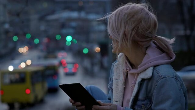 Girl With Tablet In Her Hands. Cute Young Girl With Pink Hair On The Background Of The Evening City. She Wears Hood.