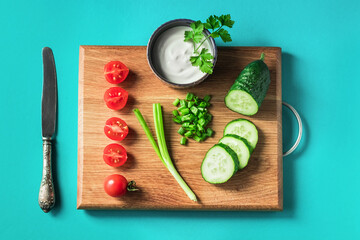 Ingredients for salad cherry tomatoes, cucumber and green onions on a cutting wooden board on a bright blue background.