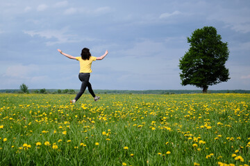 A girl in a yellow T-shirt moves across the field with flowers. Yellow flowers, green grass, blue sky, oak. Travel outside the city. Leisure
