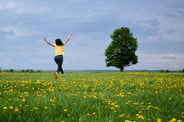 A girl in a yellow T-shirt moves across the field with flowers. Yellow flowers, green grass, blue sky, oak. Travel outside the city. Leisure
