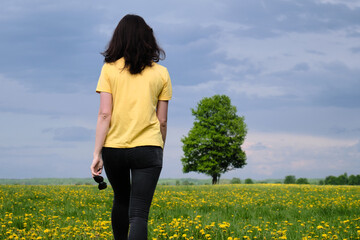 Fototapeta premium A girl in a yellow T-shirt moves across the field with flowers. Yellow flowers, green grass, blue sky, oak. Travel outside the city. Leisure