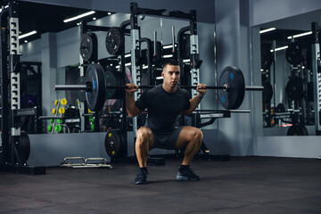 A side portrait of a bodybuilder in grey doing squats using a barbell in a gym to train his legs...