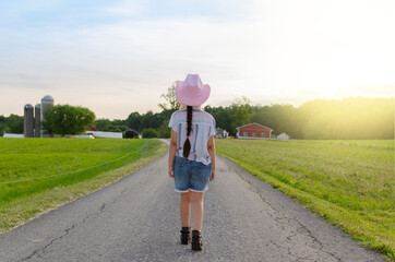 Girl walking on the road to the farm during the sunset surrounded by green grass and wearing a pink...