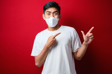 Young handsome man wearing medical mask smiling and looking at the camera pointing with one hands and fingers to the side