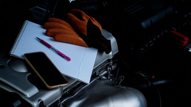 A Cell Phone, White Paper, A Pen And A Mechanic's Glove Placed On The Engine In The Car Engine Compartment, The Concept Is Self-maintenance Of The Engine Using An Smartphone App.