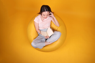 Beautiful young south east Asian woman sits on a yellow beanbag seat orange yellow color background pose fashion style elegant beauty mood expression rest relax think emotion read book top view