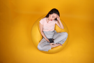 Beautiful young south east Asian woman sits on a yellow beanbag seat orange yellow color background pose fashion style elegant beauty mood expression rest relax think emotion smartphone