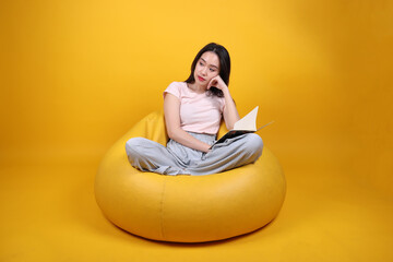 Fototapeta na wymiar Beautiful young south east Asian woman sits on a yellow beanbag seat orange yellow color background pose fashion style elegant beauty mood expression rest relax read book think