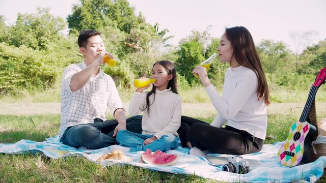 Happy family picnic. Asian parents (Father, Mother) and little girl drinking orange juice and have fun and enjoyed ourselves together during picnicking on a picnic cloth in the green garden