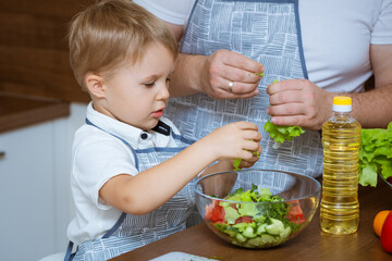 The blonde-haired son and father prepare a salad of colorful, fresh vegetables together in the kitchen. They both wore blue aprons.