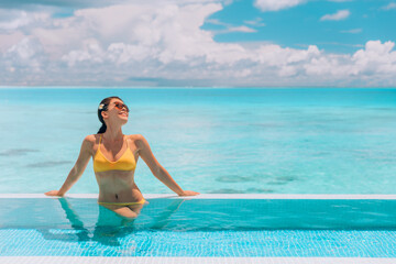 Summer sunbathing happy bikini woman swimming in infinity pool of luxury resort hotel relaxing in idyllic overwater ocean view suite. Suntan solar skin care.