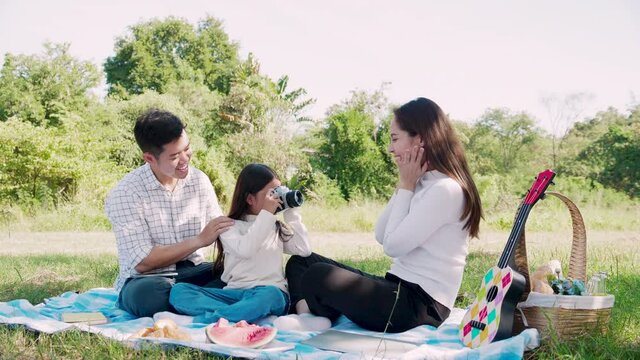 Happy family picnic. Asian parents (Father, Mother) are teaching their daughters to take pictures with film camera and have enjoyed ourselves together while picnicking on picnic cloth in green garden