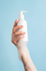 Womans hand holding cosmetics bottle with foam bubbles on pastel blue background. Bath accessories...
