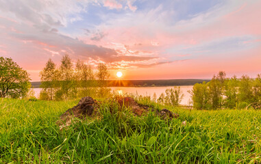 Scenic view at beautiful sunset on a shiny lake with old rough stone on the foreground, green grass, birch trees, golden sun rays, calm water ,nice cloudy sky on a background, spring landscape