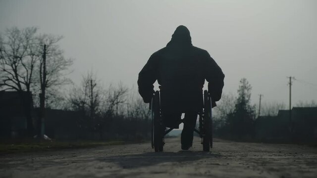 Amputee man silhouette in warm jacket rides wheelchair on ground road in grim village on cold spring evening backside view slow motion