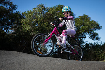 caucasian young girl child pink bike blue helmet pump bmx track riding cycle outdoors sky trees fun...