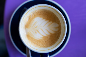 overflowing cup of latte coffee with drawn leaf on purple table, horizontal.