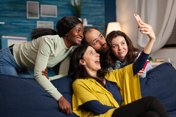 Multiethnic friends making funny faces while taking selfie photo partying in apartment, drinking...