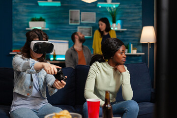 Woman and multiethnic friends playing online video games experiencing virtual reality with headset and wireless controller, having fun late in the evening sitting on couch.