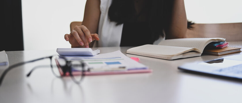Cropped shot of young woman accountant using calculator and analyzing finance data on white office desk.