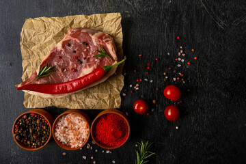 Raw piece of ribeye beef meat on on craft paper on a wooden table with tomato, basil, himalayan salt, hot peppers, paprika, spice. Fresh food. Everything for a barbecue.