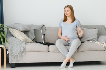 Pregnant woman with glass of water sitting on sofa. Young expectant blonde rest at home, copy space. Pregnancy, healthcare, thirst concept