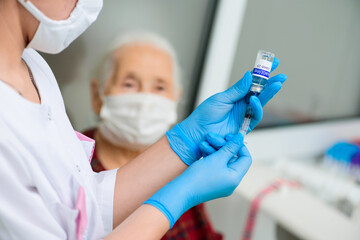 a nurse holds a syringe and a glass jar labeled covid-19 vaccine and prepares to vaccinate an elderly woman for the prevention of coronavirus infection.