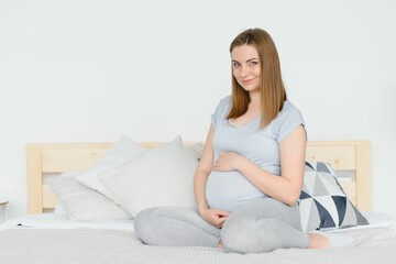 Beautiful pregnant woman holds hands on belly in bedroom at home. Young mother waiting of a baby. Concept of pregnancy, maternity, health care, gynecology, medicine. Close-up, indoors.