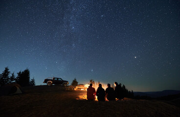 Evening starry sky over mountain valley with car and hikers near campfire. Group of travelers...