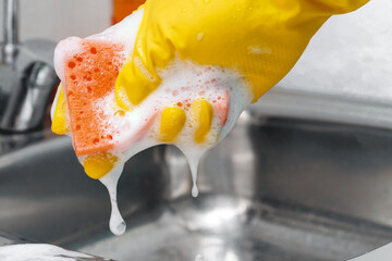 Hand in yellow latex protective glove squeezing out some foam from orange sponge.