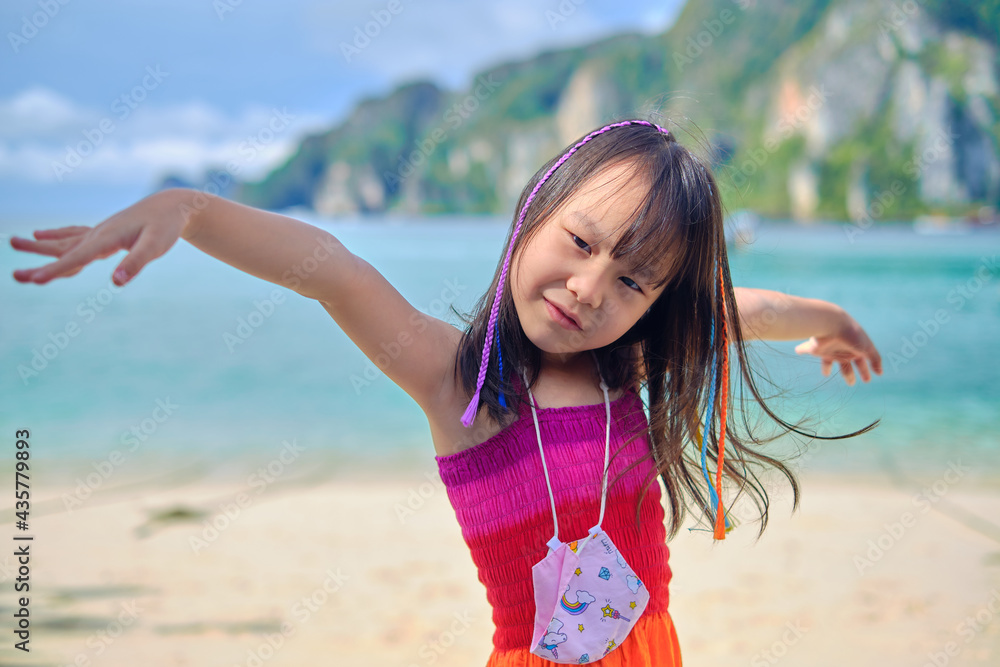 Wall mural Cute portraits of black-haired smiling Asian girl braided with multicolored thread in casual rainbow dress and face mask, beautiful beach with mountain and sea in summer holiday vacation.