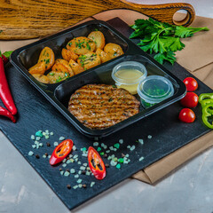 Pork Cutlet With Fried Potatoes Halves In A Plastic Box Standing On Black Stone Tray Ready For Delivery