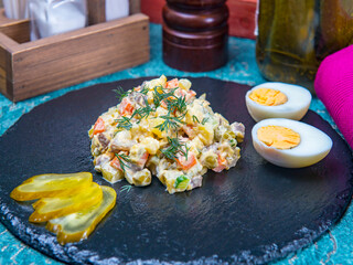 Russian salad on the round black stone tray with halves of boiled egg and pickled cucumbers aside