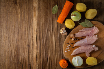 raw chicken breast fillet on a cutting board with ingredients - raw potatoes, carrots, onions, garlic and spices on a textured wooden background before cooking. top view with copy space