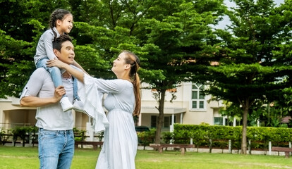 Portrait of asian happy family of three having fun together in park,   father and mother holding little daughter sitting on father back walking and having fun moments good time in summer park at home